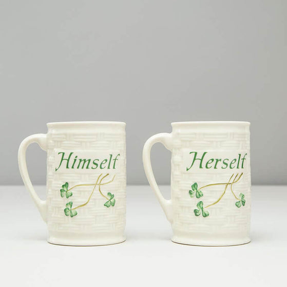 Two Belleek china mugs are presented side by side, one labeled Himself and the other Herself, both featuring green lettering and a subtle leafy design. The background is plain and neutral in color.