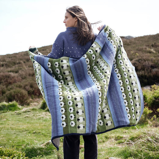 Woman holding a large, blue and green sheep patterned knitted blanket outdoors in a natural setting.