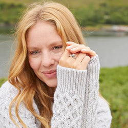 A woman with long, wavy blonde hair smiles softly while holding her hand near her face showing a silver claddagh ring on her finger. She wears a cozy light gray sweater with textured knit patterns. In the background, green grass and a lake create a serene outdoor setting.