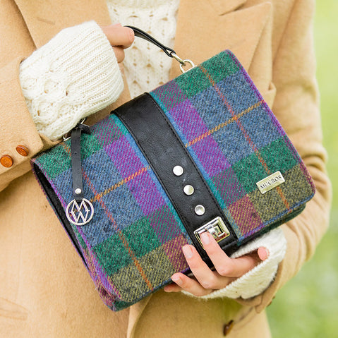 A multicolored tweed handbag with a black leather handle and metal twist clasp, sitting on a mossy surface.