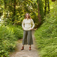 A woman stands on a path surrounded by lush greenery. She is wearing a light-colored  Aran sweater and wide-legged gray trousers, with brown shoes. The background features dense foliage and trees, creating a serene atmosphere.