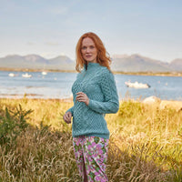 A woman with long red hair stands in a grassy field by a body of water, wearing a light blue Aran sweater and a floral skirt. The background features mountains and boats on the water, suggesting a scenic outdoor setting.