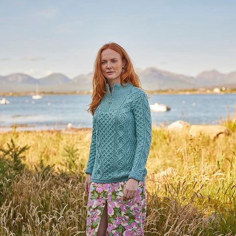 A woman with long red hair stands in a grassy field by a body of water, wearing a light blue Aran sweater and a floral skirt. The background features mountains and boats on the water, suggesting a scenic outdoor setting.