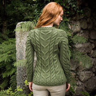 A woman with long, wavy red hair stands beside a stone wall with her back to the camera. She wears a green cable-knit Aran sweater and beige pants, posing naturally in a serene outdoor setting.