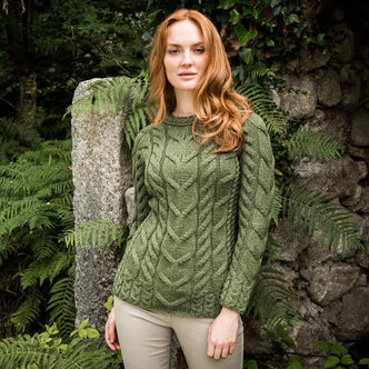 A woman with long, wavy red hair stands beside a stone wall covered in ferns. She wears a green cable-knit Aran sweater and beige pants, posing naturally in a serene outdoor setting.