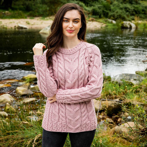 A woman with long, wavy dark hair stands beside a river and greenery. She wears a pink cable-knit Aran sweater and black pants, posing naturally in a serene outdoor setting.