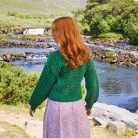 A woman with long red hair stands with her back to the camera, wearing a green knitted Aran sweater and a purple skirt. She is in a natural landscape near a river with rocks and greenery, with a waterfall visible in the background.