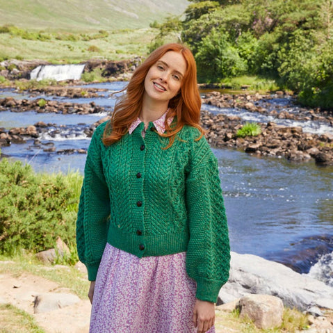 A woman stands by a river, smiling and wearing a green knitted Aran cardigan with buttons. She pairs it with a pink floral skirt, and the backdrop features greenery
