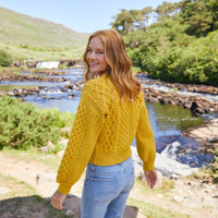A woman stands with her back to the camera looking over her shoulder, wearing a yellow knitted Aran sweater and light blue jeans in front of a serene river surrounded by lush greenery and rocky terrain. The sunlight illuminates the scene, enhancing the natural landscape.