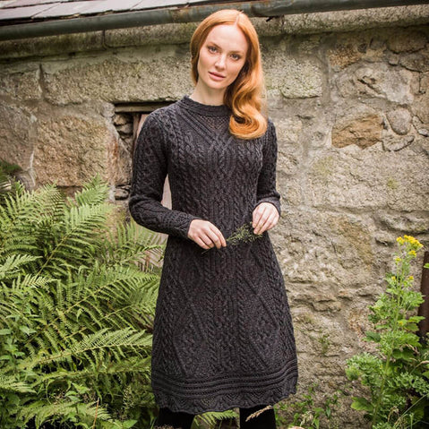 A woman stands near a stone wall, wearing a long-sleeved, dark Aran sweater dress with a textured pattern. She has red hair and islooking directly into the camera. In the background, there is an old wooden door partially concealed by ferns.