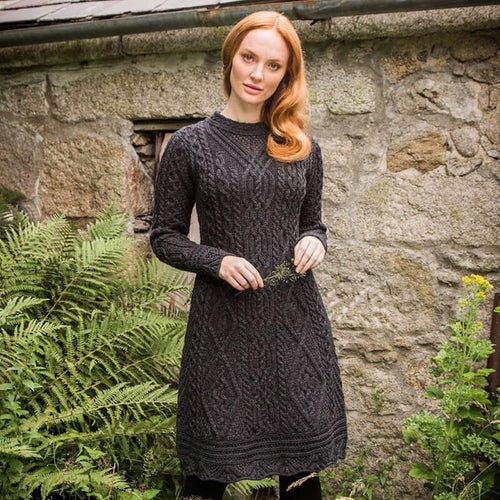 A woman stands near a stone wall, wearing a long-sleeved, dark Aran sweater dress with a textured pattern. She has red hair and islooking directly into the camera. In the background, there is an old wooden door partially concealed by ferns.