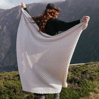 Image featuring the Traditional Aran Honeycomb Wool Throw in cream, crafted from premium wool with authentic Irish honeycomb knit patterns, wrapped around a person against a breathtaking coastal landscape, showcasing timeless Aran craftsmanship and heritage-inspired design.