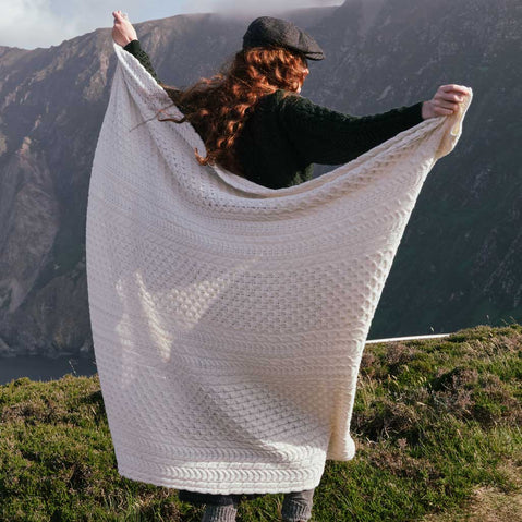 Image featuring the Traditional Aran Honeycomb Wool Throw in cream, crafted from premium wool with authentic Irish honeycomb knit patterns, wrapped around a person against a breathtaking coastal landscape, showcasing timeless Aran craftsmanship and heritage-inspired design.