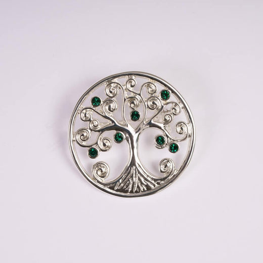 A silver brooch with the tree of life design, featuring swirling branches and roots. The tree is adorned with small green gemstones, all set within a circular border. The background is plain white.