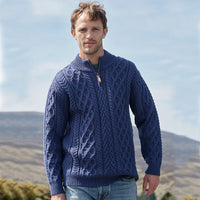 Man wearing a blue Men's Signature Half Zip Irish Wool Sweater, featuring intricate cable knit patterns and a half-zip collar, standing outdoors with hills and a partly cloudy sky in the background.
