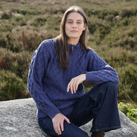 Person wearing a blue Women's Signature Half Zip Irish Wool Sweater, featuring intricate cable knit patterns and a half-zip collar, sitting on a rock in an outdoor setting with greenery in the background