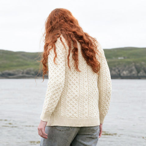 Back view of The Banna Aran Cardigan in cream, crafted with authentic Irish Aran cable knit patterns and timeless heritage-inspired styling, showcasing the intricate knit design against a scenic coastal landscape inspired by Ireland's rugged countryside.