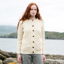 Image featuring The Banna Aran Cardigan in cream, crafted with authentic Irish Aran cable knit patterns, a timeless button-front silhouette, and practical front patch pockets, showcasing traditional Irish craftsmanship against a scenic coastal landscape.