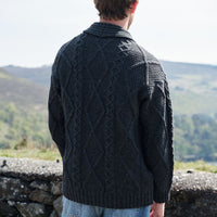 Person wearing a dark knitted sweater with a pattern, standing against a scenic background of mountains and greenery.