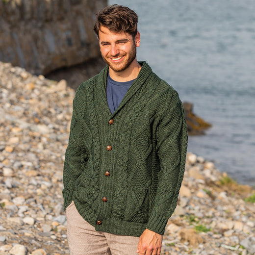 Man wearing a green Men's Heritage Aran Shawl Collar Button Cardigan standing on a pebbly beach with water in the background
