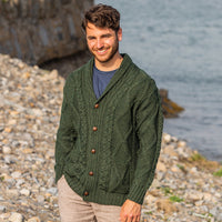 Man wearing a green Men's Heritage Aran Shawl Collar Button Cardigan standing on a pebbly beach with water in the background