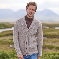 Man wearing an oatmeal Men's Heritage Aran Shawl Collar Button Cardigan in a scenic outdoor setting with mountains and fields.
