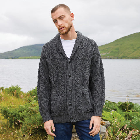 Man wearing a charcoal Men's Heritage Aran Shawl Collar Button Cardigan standing by a lake with mountains in the background