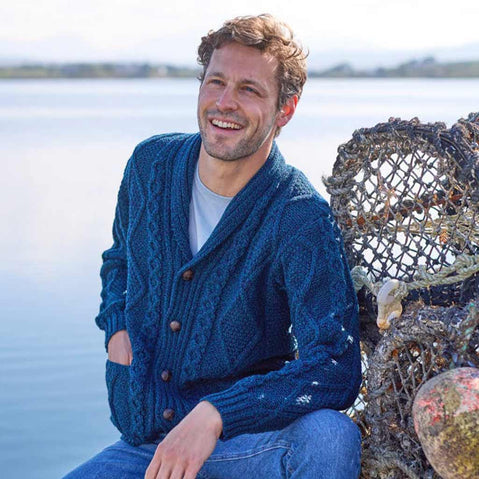 Man wearing a blue Men's Heritage Aran Shawl Collar Button Cardigan sitting by a body of water with fishing nets in the background.