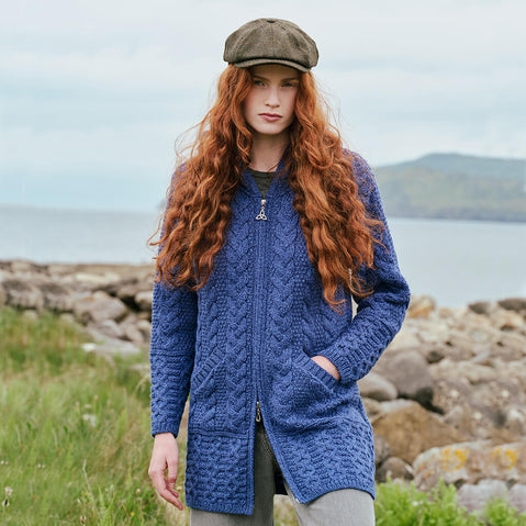 Image featuring the Hooded Aran Zipper Classic Coat in blue, crafted with authentic Irish Aran cable knit patterns, a practical hood, full-zip closure, and spacious front pockets, showcasing timeless Irish craftsmanship against a scenic coastal landscape.