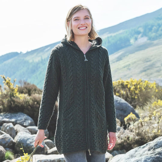 Woman wearing a classic Irish Aran hooded zipper coat in army green, standing outdoors with mountain scenery