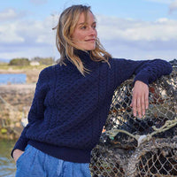 Woman wearing a navy blue Women's Signature Fisherman Turtleneck Wool Sweater standing next to lobster traps with a body of water and sky in the background.