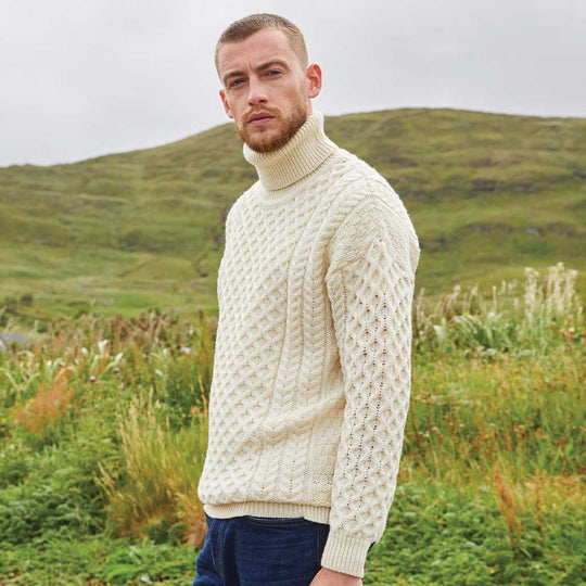 Man wearing a cream Men's Signature Fisherman Turtleneck Wool Sweater standing in a grassy field with hills in the background