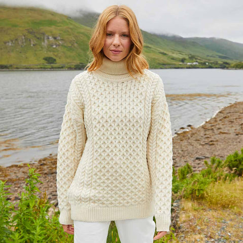 Woman wearing a cream Women's Signature Fisherman Turtleneck Wool Sweater standing by a lake with mountains in the background