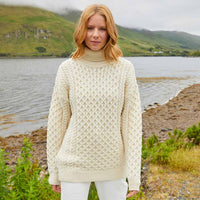 Woman wearing a cream Women's Signature Fisherman Turtleneck Wool Sweater standing by a lake with mountains in the background