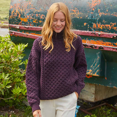 Woman wearing a damson Women's Signature Fisherman Turtleneck Wool Sweater standing in front of a rustic green boat.
