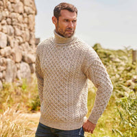 Man wearing an oatmeal Men's Signature Fisherman Turtleneck Wool Sweater standing in a natural setting with stone wall and greenery.