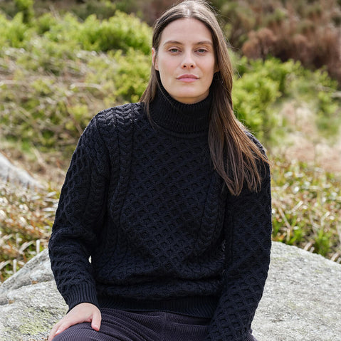 Woman wearing a black Aran sweater sitting on a rock with greenery in the background