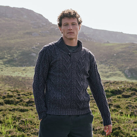 Image featuring the Men's Classic V-Neck One Button Aran Sweater in cormorant blue, crafted with authentic Irish Aran cable knit patterns and a signature one-button shawl collar, showcasing timeless Irish craftsmanship in a scenic countryside setting.