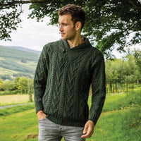 Man wearing a  green V-neck Irish wool Aran sweater from The Irish Store. He stands outdoors amid lush greenery with hills in the background.