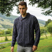 Man wearing a dark blue knitted Aran sweater standing in a scenic outdoor setting with greenery and mountains.