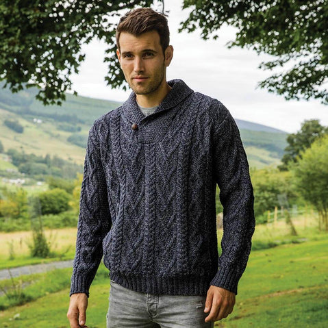 Man wearing a dark blue knitted Aran sweater standing in a scenic outdoor setting with greenery and mountains.