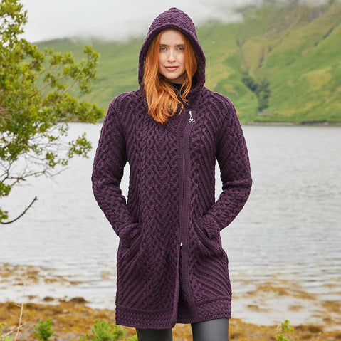 Woman wearing a damson Women's Heritage Aran Hooded Zipper Coat standing by a lake with green hills in the background