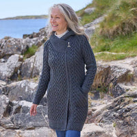 Woman wearing a charcoal Women's Heritage Aran Hooded Zipper Coat standing on rocky terrain with a body of water and grass in the background.