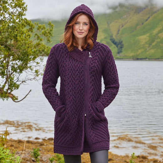 Image featuring the Women's Heritage Aran Hooded Zipper Coat in damson, crafted with authentic Irish Aran cable knit patterns, a practical hood, full-zip closure, and spacious front pockets, showcasing timeless Irish craftsmanship against a scenic lakeside landscape inspired by Ireland's rugged countryside.
