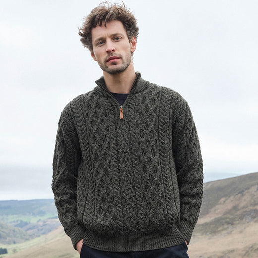 Man wearing an army green cable knit Aran sweater standing in a scenic outdoor setting with hills and a cloudy sky.