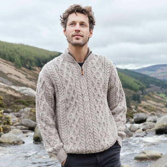 Man wearing a cable knit beige Aran sweater standing by a river with mountains in the background