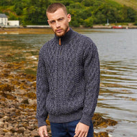Man wearing a dark blue knitted Aran sweater standing by a body of water with greenery in the background