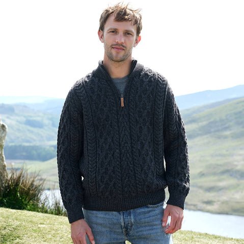 Man wearing a charcoal gray Aran sweater standing outdoors with a scenic background