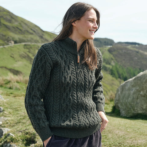 Woman wearing a green cable knit sweater in a scenic outdoor setting with hills and rocks.