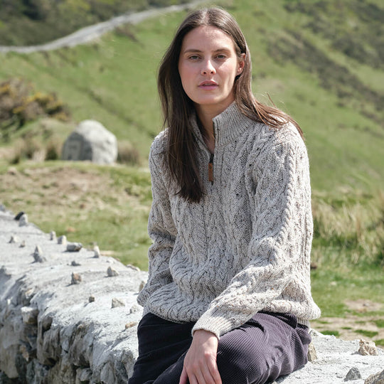 Woman wearing a cable knit sweater sitting on a stone wall with a scenic background